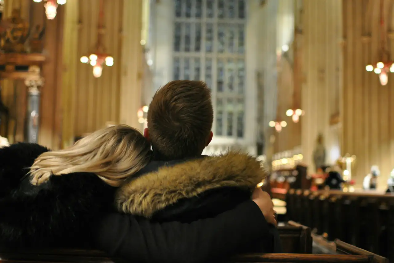 A man and a woman, seen from behind, sitting side by side in a wooden church pew, facing the altar.